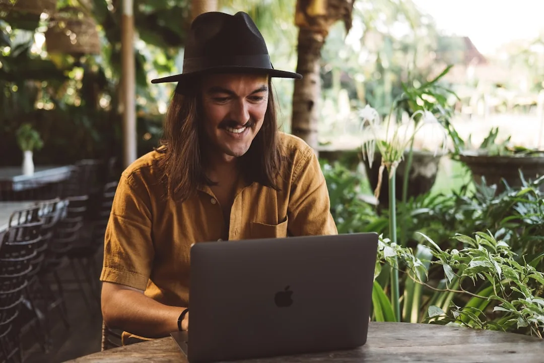Man in black hat smiling at laptop in a lush tropical garden café