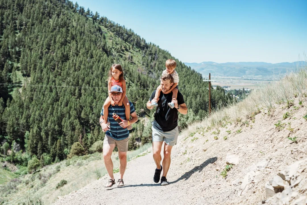 Two fathers carrying their children on shoulders on a mountain hiking trail