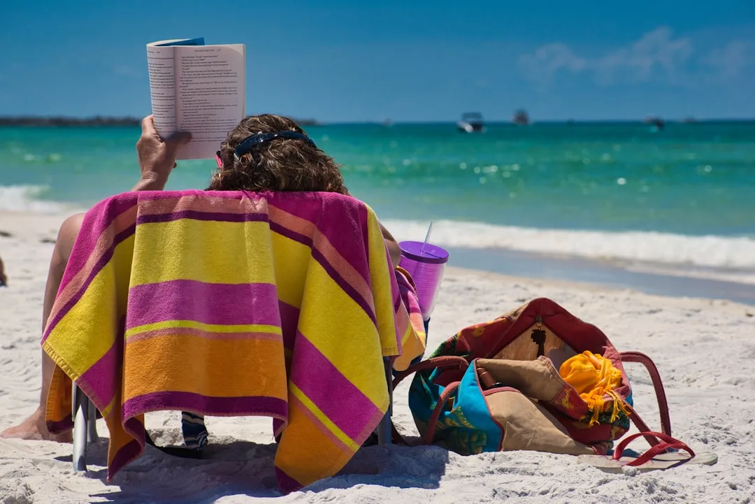 Person mit buntem Strandtuch lesend am türkisfarbenen Meer – entspannter Strandtag