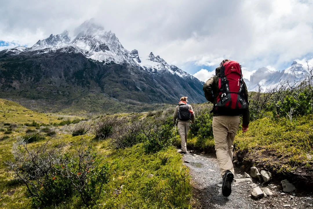 Two hikers with trekking backpacks on a mountain trail with snow-capped peaks in the background